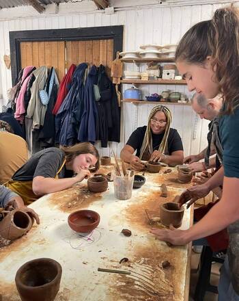 Four people shape brown clay pots at a messy art studio table. A woman with blonde braids smiles, another concentrates.