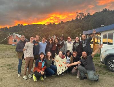 A smiling group of 17 people pose outside with a vibrant orange sunset illuminating the sky over forested hills and cabins.